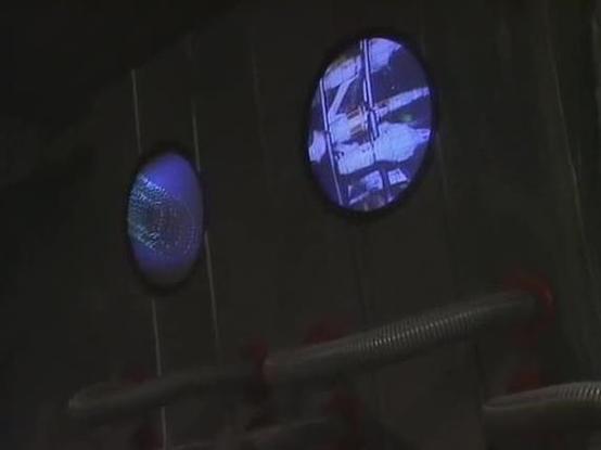 Claude Opus 4.6 describes the image as: "This image is from the classic BBC science fiction series **Blake's 7**. The scene shows part of the interior set design characteristic of the show, featuring a dark, industrial-looking wall with two circular viewscreens or monitor portals. The left circular display emits a blue glow with what appears to be a textured or grid-like pattern, while the right circular screen displays what looks like a video feed showing some kind of spacecraft or station ima…