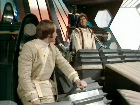 Claude Haiku 4.5 20251001 describes the image as: "# Blake's 7 Scene Description

This image captures a scene from the classic BBC Television series "Blake's 7," set within the futuristic interior of a spacecraft. The setting features sleek, metallic corridors with geometric paneling and industrial design elements typical of the show's distinctive aesthetic. Hexagonal light panels illuminate the space, and various technological apparatus line the walls, creating an atmosphere of advanced space …