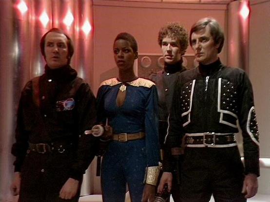 Claude Sonnet 4.5 20250929 describes the image as: "This image shows four crew members standing in a corridor with distinctive pink-tinted lighting and diamond-shaped wall fixtures. The scene appears to be aboard a spacecraft, with the characteristic production design of the BBC series.

From left to right: Michael Keating wears a black uniform with a high collar and what appears to be a communication device on his chest. Josette Simon stands in a striking blue outfit with ornate gold shoulder …