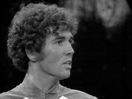 Claude Opus 4.6 describes the image as: "This black-and-white image shows actor Steven Pacey in his role as Del Tarrant from the classic BBC Television series "Blake's 7." Pacey appears young here, with his distinctive curly hair, captured in profile view facing to the right. He is wearing what appears to be a light-colored, high-collared jacket with a zipper, consistent with the futuristic costume design typical of the series. The photograph has a dramatic quality, with Pacey's expression sugg…