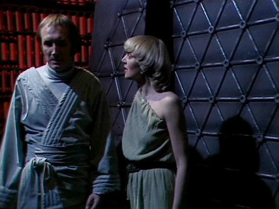 Claude Opus 4.6 describes the image as: "This image is from the classic BBC Television series "Blake's 7." The scene features Michael Keating, who plays the lovable thief Vila Restal, standing alongside Carol Hawkins, who appears as a guest character in the series.

Keating is dressed in Vila's characteristic costume — a light blue-grey tunic with a high collar and belted waist, fitting the show's distinctive sci-fi aesthetic. He has a somewhat wary or concerned expression, typical of Vila's ca…