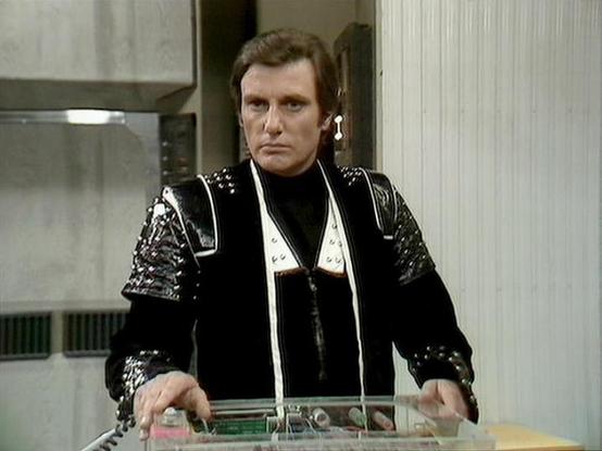 Claude Haiku 4.5 20251001 describes the image as: "# Blake's 7 Scene

This image captures a scene from the classic BBC science fiction series "Blake's 7." The actor portrays Kerr Avon, the cunning and ambitious computer expert of the rebel crew. He is dressed in his characteristic black outfit adorned with elaborate silver studs and embellishments across the shoulders and sleeves, reflecting the futuristic aesthetic of the series. A white scarf drapes elegantly around his neck, adding to his di…