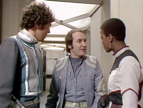 Claude Haiku 4.5 20251001 describes the image as: "# Blake's 7 Scene Description

This image captures a tense moment aboard a spacecraft corridor in the classic BBC series Blake's 7. Three crew members are engaged in conversation within a utilitarian sci-fi setting, characterized by the distinctive 1970s aesthetic of the show's production design.

The corridor features the series' signature metallic panels, geometric lighting fixtures set into the ceiling, and a minimalist interior that emphasi…