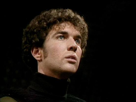 Claude Sonnet 4.6 describes the image as: "This image appears to be from the classic BBC Television series **Blake's 7**, likely captured during a dramatic scene. The setting is dark and atmospheric, with a nearly black background that emphasizes the subject's face through stark, focused lighting — a hallmark of the show's stylized production aesthetic.

The young man in the frame has curly dark brown hair, striking light-colored eyes, and is wearing a dark turtleneck or high-collared costume t…