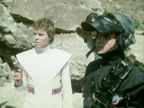 Claude Haiku 4.5 20251001 describes the image as: "# Blake's 7 Scene

This image shows two characters in a scene from the classic BBC Television series "Blake's 7." The character on the left wears a distinctive white tunic with purple trim, while the figure on the right is dressed in dark military-style armor and helmet, suggesting a confrontational encounter between characters of different allegiances.

The rocky, concrete setting appears to be an industrial or underground location, typical of…