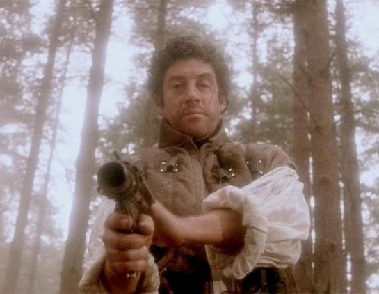 Claude Opus 4.6 describes the image as: "This image shows Gareth Thomas in his iconic role as Roj Blake from the classic BBC Television series *Blake's 7*. He is standing in a misty, wooded setting surrounded by tall trees, pointing what appears to be a flintlock-style pistol directly at the camera. Thomas is dressed in period-style costume — a brown leather jerkin or vest over a white billowy shirt — which suggests this scene takes place in a historical or rustic setting, possibly on a primiti…