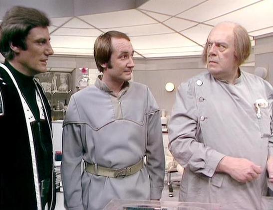 Claude Opus 4.6 describes the image as: "This image is from the classic BBC Television series *Blake's 7*. The scene features three men in what appears to be a futuristic interior setting, consistent with the show's science fiction aesthetic.

On the left is Paul Darrow as Kerr Avon, the cold and calculating computer expert, wearing his characteristic dark clothing. His sharp features and intense gaze are trademark Avon.

In the center is Michael Keating as Vila Restal, the lovable, cowardly th…