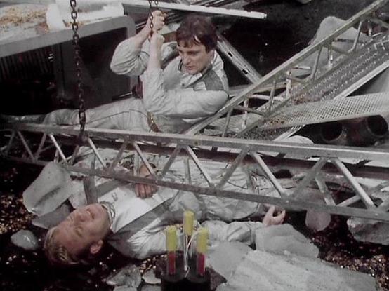 Claude Haiku 4.5 20251001 describes the image as: "# Blake's 7 Scene Description

This image captures a dramatic moment from the classic BBC Television series "Blake's 7," showing two characters in a precarious situation involving industrial scaffolding and chains. One actor is positioned higher on the metal framework, while another lies below on the ground, creating tension in the scene.

The setting appears to be an industrial or mechanical environment, with metal girders and structural eleme…