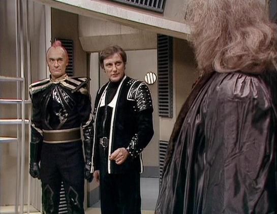 Claude Haiku 4.5 20251001 describes the image as: "# Blake's 7 Scene

This image captures a tense moment from the classic BBC science fiction series "Blake's 7." The scene is set in what appears to be a spacecraft corridor or interior chamber, featuring sleek, minimalist 1970s design elements with neutral walls and functional details.

Three figures are present in the frame. On the left stands a character in elaborate black and gold military uniform with distinctive shoulder pieces and ornate d…