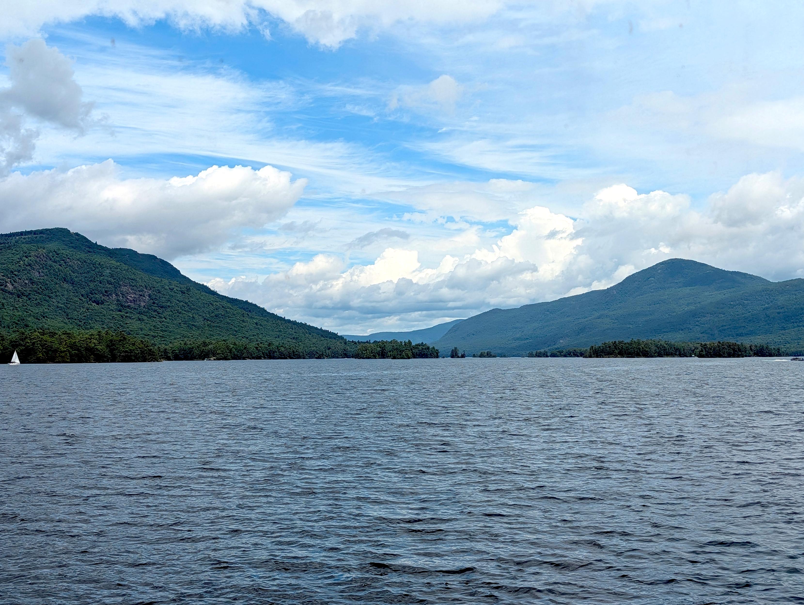 Background: Blue sky with high and low altitude clouds...
Middleground: Distant mountains and lake...
Foreground: Mountain peaks on the left and right... Green tree lined shoreline and lake water with minor waves... Distant sailboat on the left and small motorboat on the right... 

Created: 6/23/26 1:05pm
Aperture: ƒ∕1.68
Exposure: 1∕831 s
ISO: 24
Focal Length: 6.9 mm
Google Pixel 10 Pro XL