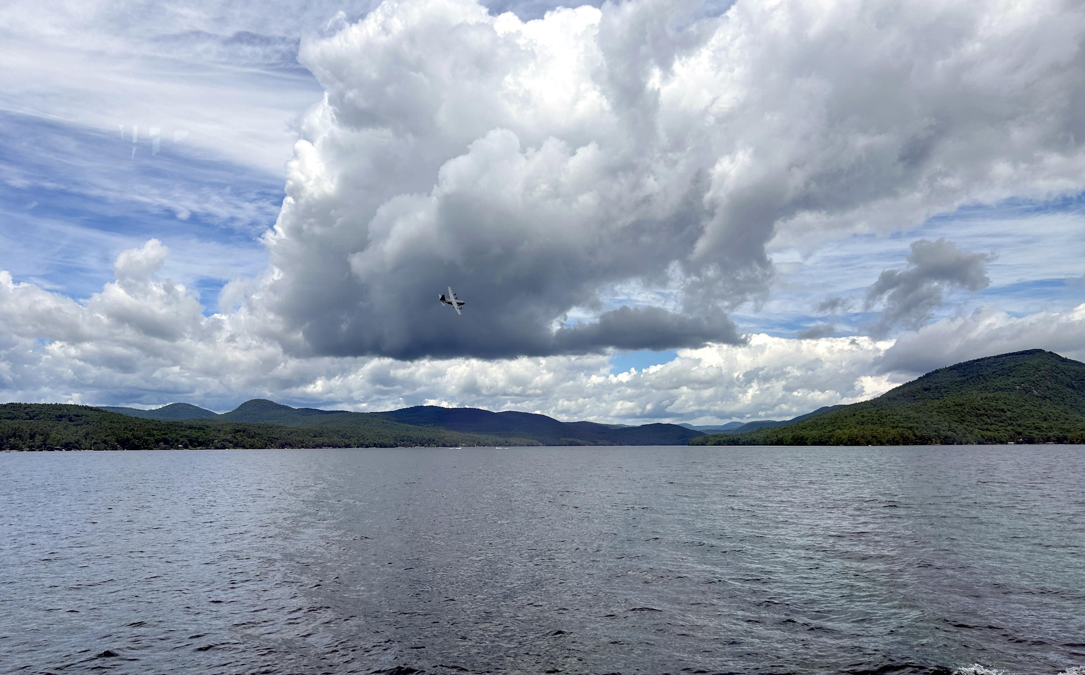 Background: Clear blue sky with high wispy clouds...
Middleground: Low level puffier cumulus clouds... Small mountains and tree lined shoreline...
Foreground: My best identification is a Lockheed C-130 Hercules (?) aircraft as it flew over during a lake boat cruise... 

Created: 8/11/26 2:25pm
Aperture: ƒ∕1.5
Exposure: 1∕3831 s
ISO: 50
Focal Length: 5.7 mm