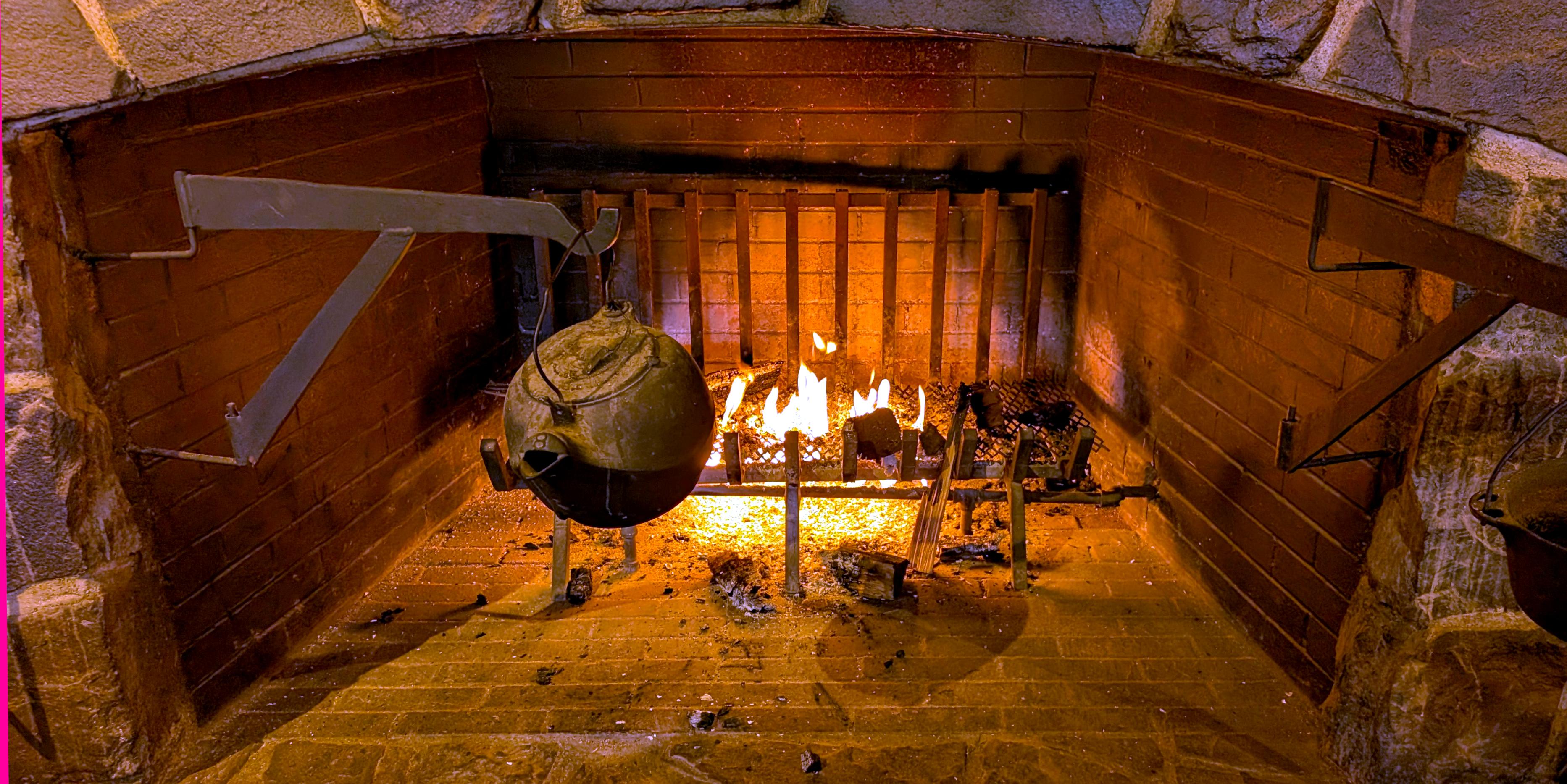 Background: Glowing back brick wall of a fireplace and a fence-like grating cast a shadow on the wall...
Middleground: Metal fireplace grating holding wooden logs glowing with fire... Some burnt log pieces have fallen to the below the grating...
Foreground: Stone fireplace walls with attached rotating cast iron hooked pot holders on the left and right holding a black kettle on the left and a black pan on the right...

Created: 2/3/2026 1:00pm
Aperture: ƒ∕1.68
Exposure: 1∕134 s
ISO: 275
Focal Length: 6.9 mm