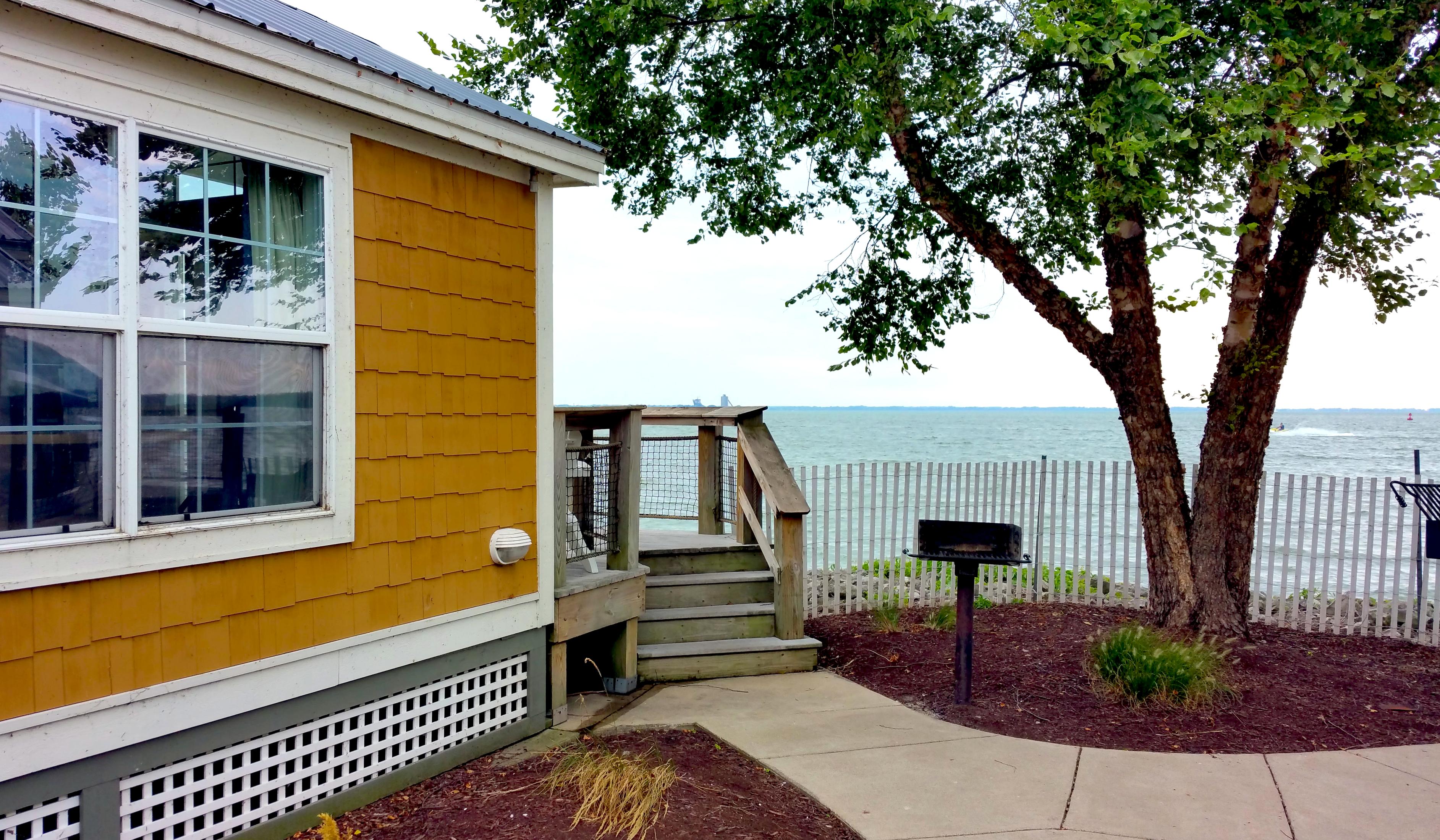 Background: Cloudy sky and barely visible land
Middleground: Lake Erie with a jet skier and a red buoy on the right
Foreground: Lakeside cottage with dark metal roof, orange shingles, window, white lattice crawl space, and wooden stairs leading to a netted fenced deck... At center, a black small barbecue and at right, a tree with green leaves with green bush and mulch base... Cement pathway and white picket fence behind tree... Nice place...

Created: 8/11/2017 3:26 PM
Aperture: ƒ∕2.2
Exposure: 1∕326 s
ISO: 40
Focal Length: 4.8 mm