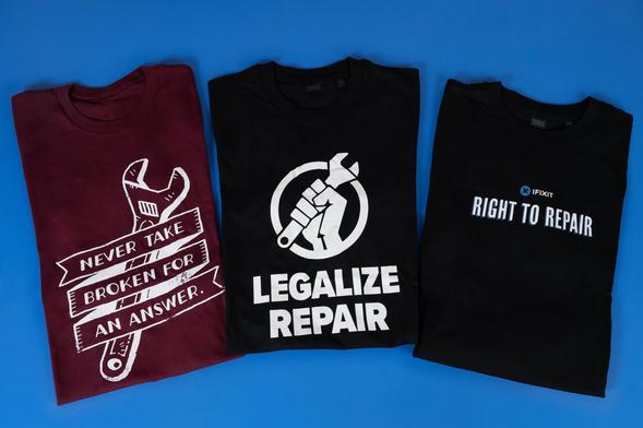 Three folded repair-themed T-shirts displayed on a blue background. A burgundy shirt features a wrench and the phrase “Never take broken for an answer,” while two black shirts read “Legalize Repair” and “Right to Repair” alongside iFixit branding.
