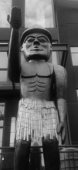 Friendship welcome totem pole at Stó:lõ First Nations reserve office. 2022. The totem is of a Stó:lõ Indigenous man with right hand extended out. Wearing a weaved skirt & a cone shaped hat. It stands in front of an office building.
Black and white photo.