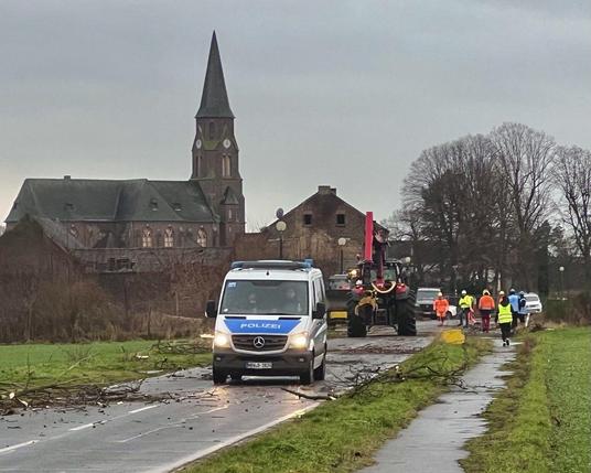 Foto von Straße mit teils gerodeten Bäumen. Vorn ein Polizeiwagen, dahinter Trecker mit Kettensägen-Schwenkarm und Menschen mit Leuchtkleidung.

Äste liegen auf der Straße. im Hintergrund ist die entweihte Kirche von Manheim mit vernagelten Fenstern zu sehen.