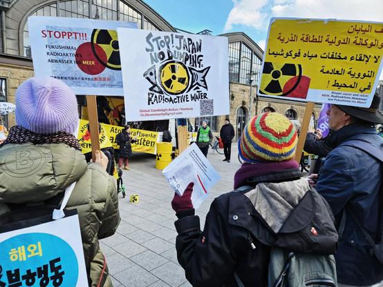 Demonstrierende auf dem Platz vor dem Hamburger Hbf. Sie halten Schilder hoch, in der Mitte "Stop Japan No dumping radioaktive Walter!" zwischen dem Text ein Fisch mit radioaktive Zeichen. Im Hintergrund eine Rednerin vor einem Transparent und einem Atomfass. Dahinter das Hbf. Gebäude.