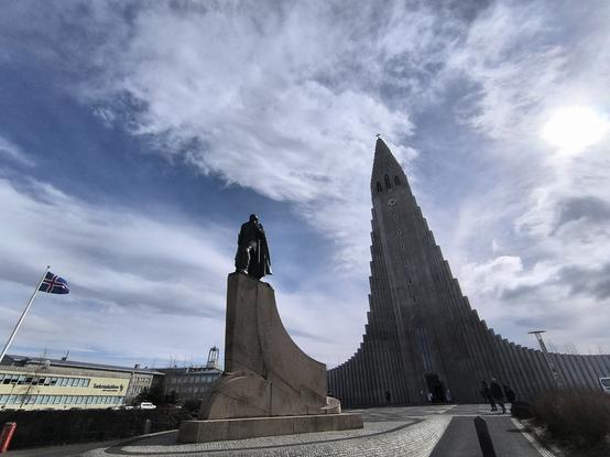 Une statue d'un viking devant une grande église. Le soleil est masqué par un voile nuageux. À gauche flotte le drapeau de l'Islande. Le bâtiment au fond est le Taekniskolinn, un centre d'apprentissage dans plusieurs métiers : peintre, menuisier, plombier ou encore électricien.