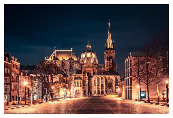Nighttime city street scene with Aachen Cathedral glowing in the background, highlighting its architectural beauty.