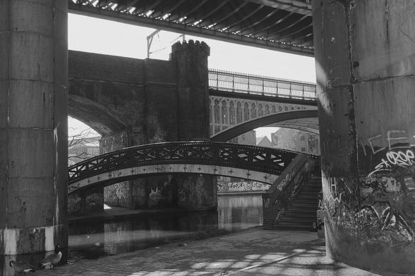 This black-and-white photograph captures an urban scene dominated by a series of bridges and architectural structures. In the foreground, a large concrete pillar stands to the left, while another pillar with graffiti is visible to the right. Between these pillars, a metal bridge with intricate designs arches over a calm canal. Above this bridge, another larger bridge with a metal railing spans the scene. The background features more bridges and industrial elements, with a staircase leading up to one of the bridges. The overall atmosphere is one of layered urban complexity