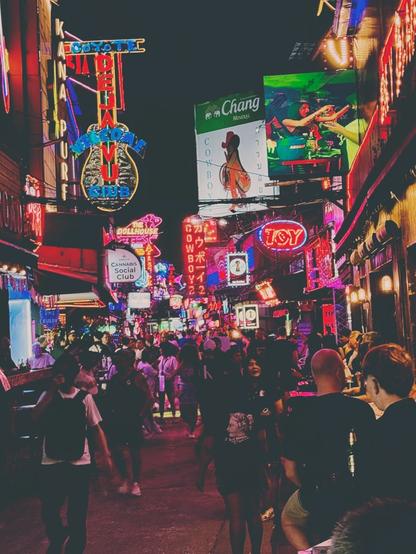 Night scene of a busy alleyway in Bangkok. Numerous neon signs in Thai, English and Japanese covering most of the black night sky, advertising for bars, cannabis shops and strip clubs. There’s a billboard for Chang beer and a large LED screen showing ads. The crowd below seems to melt together into a single mass, the further you look down the alley.