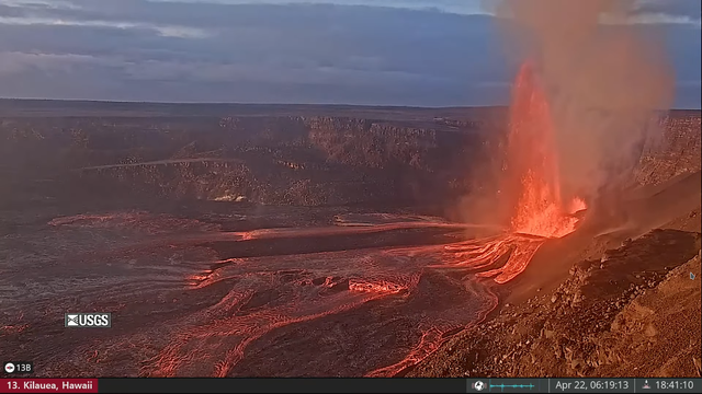 Screenshot of the 22th volcanic eruption of Kilauea 2025. The eruption is happening inside the bigger older crater. Lava is flowing to the left fanning out into the crater floor.Fountains of lava eject from the little crater. In the background the sky is colored in blues from dawn.