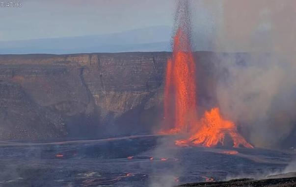 Red lava fountains erupting from a vent in a floor of dark lava, against the background of a brown crater wall and a light blue sky.