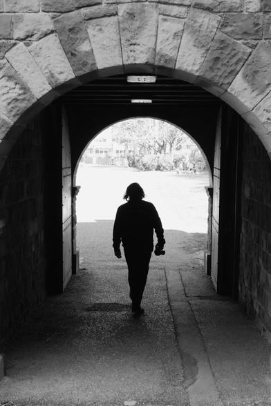 This black-and-white photograph captures a solitary figure walking through a stone archway. The archway, made of large, rough-hewn stones, frames the scene and creates a sense of depth. The person, seen from behind, is walking away from the camera, carrying a bag in one hand. Beyond the archway, a bright, open space reveals a park or garden with trees and distant buildings, creating a stark contrast with the darker tones of the archway. The overall mood is contemplative and slightly mysterious, with the silhouette of the figure adding a sense of solitude.