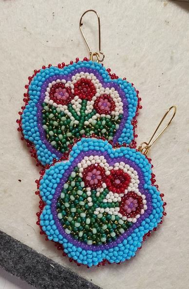 a pair of beaded earrings by kia of flowers with a light blue border, a purple outline, and red and pink stamens, filled with many colors of green and cream colored beads.