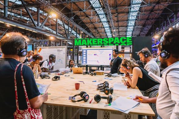 Das Bild zeigt eine Gruppe von Personen mit Kopfhörern um einen großen Holztisch. Im Hintergrund befindet sich ein Screen mit der Aufschrift „Makerspace“.