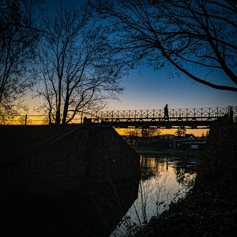 Eine Person geht über eine Brücke, links und rechts befinden sich kahle Bäume. Die gesamte Szene rie findet vor einem Sonnenuntergang statt.