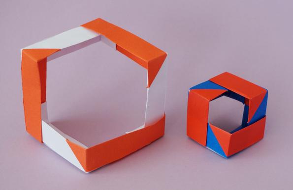 Two origami cubes on a lilac background. Each is made from six units.
The cube on the right has two opposite vertices missing, as well as parts of the adjacent faces. The missing parts of the adjacent faces are larger on the cube on the left.
The colours of the left cube are red and white; the right cube is red and blue.