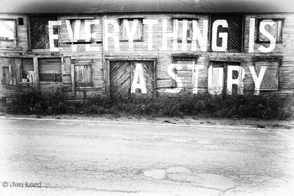 A Simple Rural Street Photo In Landscape Format. In The Foreground Is A Rough Tarmac Road Stretching From The Left To The Right And Rising Up About One Third In The Frame. On The Far Side Of This Narrow Street Is A Single Line Painted On The Side Of The Road. On The Verge Is A Strip Of Rough Undergrowth Growing About A Metre In Height. Beyond And Again Stretching From The Left To The Right Margins Is A Decaying Wooden Barn Or Warehouse. It Has A Ground And First Floor And A Pitched Felt Roof On Top. The Front Consists Of Multiple Horizontal Wooden Planks Interrupted By A Number Of Doors And Boarded Up Windows. Written Across The Building In Large White Bold Letters, Perhaps One And A Half Metres Tall, Are The Words: 'EVERYTHING IS A STORY' ('Everything Is' - On The First Floor And 'A Story' - Underneath On The Ground Floor). 
The Location: Vardø, Norway's Most Eastern Town: 31º East, Further East Than St Petersburg And Istanbul. At 70º North Its Somewhat North Of The Arctic Circle (2017).