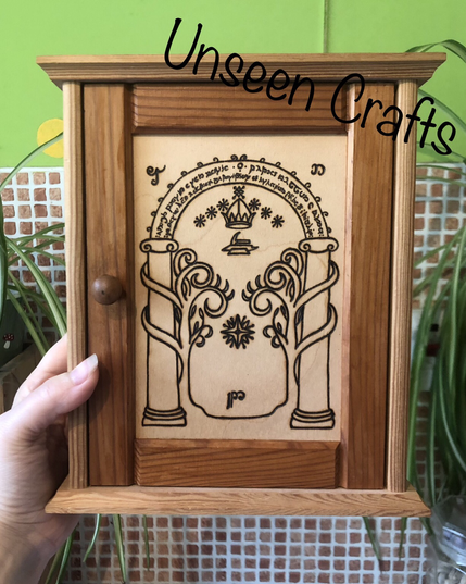 Small wooden cupboard with wooden handle. The inner frame has a pine stain and the lighter wooden middle as a burned design showing the Doors of Durin - two pillars with trees growing around, an archway above the pillars to connect them. The arch has words in elvish.