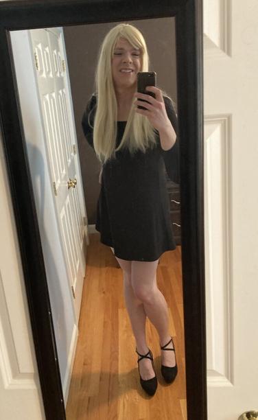mirror selfie of blonde woman with thigh length black dress and black strap high heels. smiling.