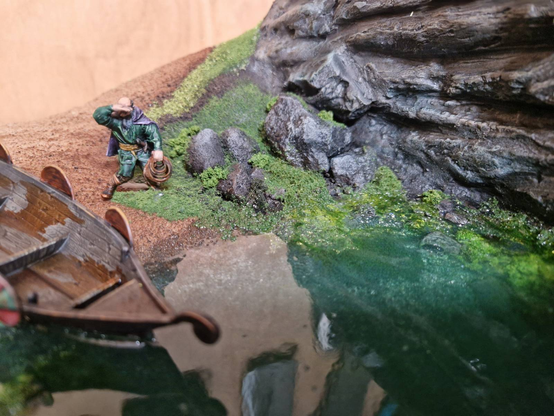 A close-up of the previous resin pour and diorama, focusing on the beach, seaweed, front of the drakkar, and the little Viking fella looking out to sea. The cliffs in the background are currently a mixture of greys, and need more colour and white picking out on them. The seaweed looks good, wet from the resin but with very tiny bubbles that make it look silvery. The resin itself is a deep, semi-transparent green-blue-grey. Clear enough that you can see the seaweed beneath it, as well as the sand on the surface of the beach.