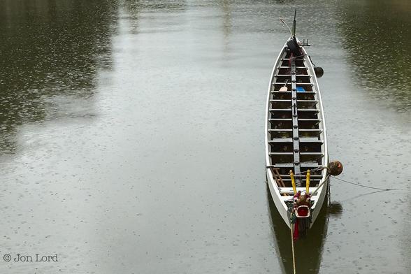 A Simple Low Contrast Colour Photo In Landscape Format. Nearly All The Image Is Filled With The Still Waters Of A River. Neither Bank Is In View. The Central Area Stretching From The Top To The Bottom Has A Wide Bright Strip Of Reflected Light From The Overcast Sky. The Left And Right Edges Have A Dark Green Tint. The Entire Surface Of The River Has Round Splash Marks From Falling Rain. To The Right Of Centre And Aligned With The Camera Is A Long Dragon Boat. It Is Moored Bow Facing The Camera And Only A Short Distance Away. Forward Of The Bow Is The Bowsprit In The Form Of A Colourful Dragon, Jaws And Eyes Wide Open. Aft Are Fourteen Positions For Her Crew With A Rowlock On The Sten For A Paddle/Tiller In Order To Steer. 
The Location: The River Silver In Mui Wo, Hong Kong (2016).