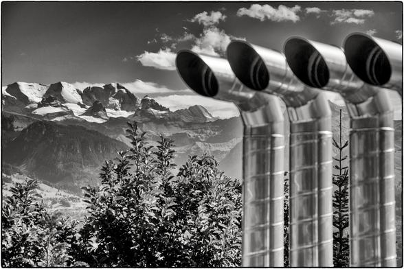 Schwarzweissfotografie zum Thema "Klimakatastrophe": links im Bild sind schneebedeckte Berge, rechts vier Abluftrohe aus Metall.