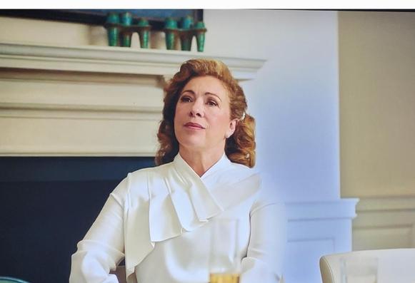 Alex Kingston plays the US Ambassador. Shown here in her residence seated at a desk. Fireplace behind her
 She is wearing a stylish white dress top. She has light brown hair and an imperious expression on her face. Chin raised.