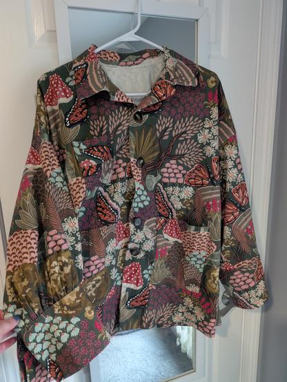 A boxy, oversized jacket hanging in front of a white door. The jacket has a collar, large cuffs at the wrists, tortoiseshell colored buttons on the front and three patch pockets. Two of the pockets are on the bottom and one is on the chest. The jacket has dropped shoulders and the sleeves are gathered at the back.

The fabric is army green with large print of botanicals in other shades of green; orange butterflies; red mushrooms; pink, blue and white flowers; and white snails, ravens, and bears.