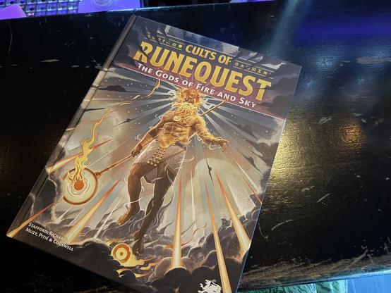 Photo of a book on a bar

On the cover there is a brightly glowing figure of an imposing bearded, crowned man, staff with fiery ball in hand. He is floating in the air, the sun rune at his feet. He is Yelm, the sun god of the game world Gloranth. 

CULTS OF RUNEQUEST
THE GODS OF FIRE AND SKY
STAFFORD, RICHARD
MUZY, PITIÉ & COGSWELL