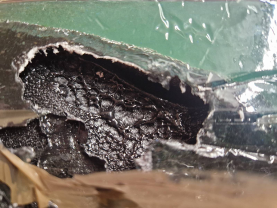 A close-up of some coloured polyester resin on top of an XPS foam base. The resin is a greenish-grey and the foam is black. The foam has melted into bubbly pockets where the heat from the curing resin has reached it.