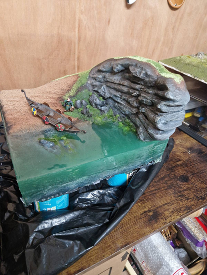 A diorama featuring a beach, a cliff, a small boat, some seaweed, and a deep block of green-blue-grey resin to mimic the sea, The front of the diorama has minimal damage from the resin curing process. Overall, it looks pretty good. Just needs some cleaning up and the diorama finishing.