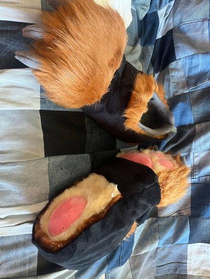 A photo of two fursuit footpaw shoes made of faux fur and minky fabric, foam, crafted around a pair of shoes. It shows lightbrown fur at the bottom, and darker brown fur everywhere else. It has pink pawpads, and a kind of "socks" around them made of black minky fabric.