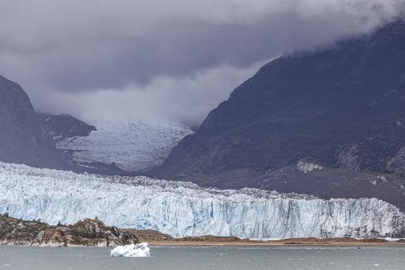 A moody, overcast view with thick, grey cloud low down and smothering the scene. We're on water at the end of a fjord looking to land where a wide U-shaped valley reaches down to the shore. The rough, rocky sides of the valley are dotted with dark bushes and vegetation almost as black as the rock they're on. Stretching across the width of the shoreline there is a glacier, dozens of metres high, roughly cracked in vertical jags of blue piercing the white ice. The glacier rises and steps back through the valley and over a small, dark rise it disappears high into the valley until it reaches the cloud that covers its uppermost reaches. A solitary berg of ice is in the flat water ahead of the shallow, pale golden shore that forms a narrow border between the fjord and glacier.