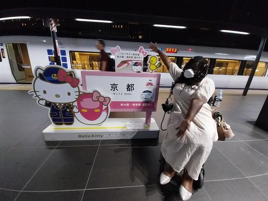 Jay sitting in her wheelchair wearing a long white dress and silver shoes on the train platform in Kyoto Station next to a large sign with Hello Kitty wearing a station master uniform and hat with her signature red bow, next to a chibi pink version of the Shinkansen train that is also wearing a bow, and text advertising the train, in front of the Hello Kitty Shinkansen itself