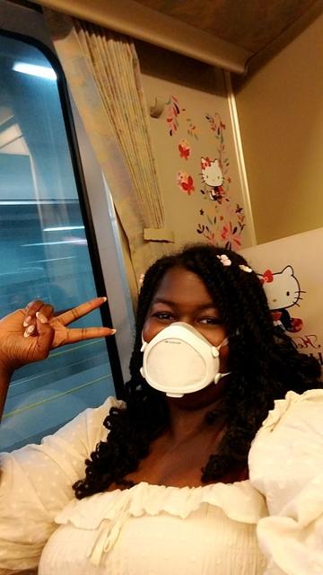 Jay sitting in her seat on the train in front of a wall with Hello Kitty themed artwork on it and a custom headrest cover that shows Hello Kitty kneeling in a blue floral kimono with the text "Hello Kitty Haruka"