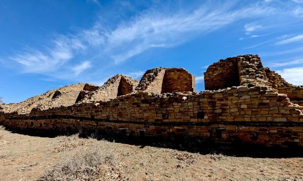 An ancient puebloan ruin: a stone wall with a linear series of fairly fall (maybe 2-story?) fairly large rooms.behind it. The wall looks like it's maybe 5 feet fall on average, and the walls of the rooms we see behind it are about twice that height. The sky is blue with whispy cirrus clouds.