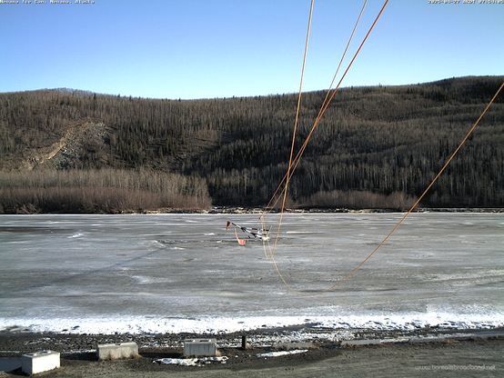 Photo from 754am AKDT Sunday, April 27 from the dock at Nenana looking across the Tanana River with the tipped over tripod still connected to the ropes to the clock tower. A ridge is seen on the far side of the river with blue skies above.