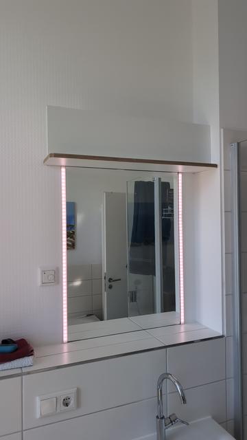 Spiegel über dem Waschbecken mit einer gefliesten Konsole. Der Spiegel hat links und rechts zwei Aluprofile mit weißen Diffusor, in dem APA101 kompatible LED Stripes sind (jeweils ca 48 LED). Darüber ein Regalbrett mit Holzdekorrand. Darauf ist als Sichtschutz ein einfaches weißes Regalbrett gestellt. Dahinter ist der WLED Controller, die Kabel und das Netzteil versteckt. Man kann auch die Befestigungswinkel nicht sehen, mit dem alles an die Wand geschraubt ist.